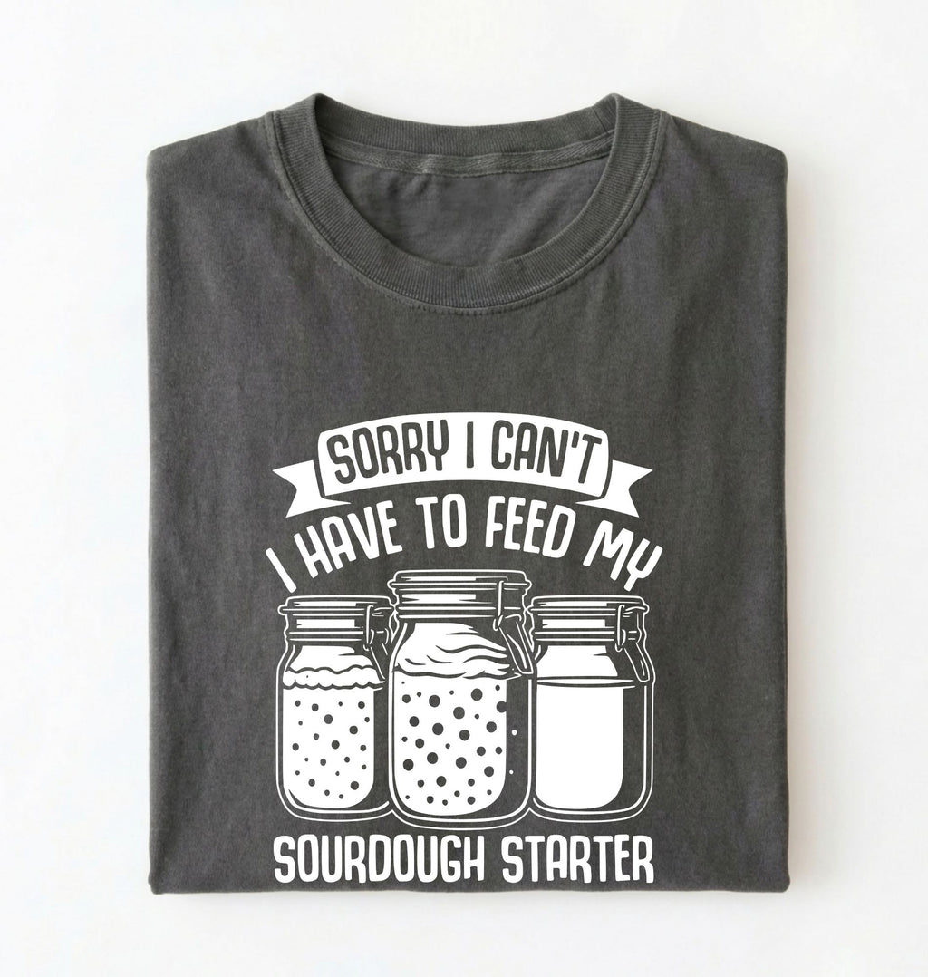 Flat lay of a pepper Sorry I Can't I Have to Feed My Sourdough Starter shirt with starter jar graphic on charcoal t-shirt on a white background 