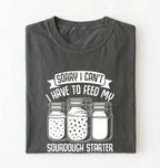 Flat lay of a pepper Sorry I Can't I Have to Feed My Sourdough Starter shirt with starter jar graphic on charcoal t-shirt on a white background 