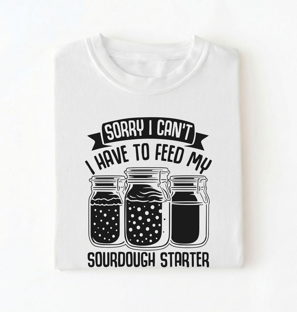 Flat lay of a white Sorry I Can't I Have to Feed My Sourdough Starter shirt with starter jar graphic on charcoal t-shirt on a white background 