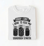 Flat lay of a white Sorry I Can't I Have to Feed My Sourdough Starter shirt with starter jar graphic on charcoal t-shirt on a white background 
