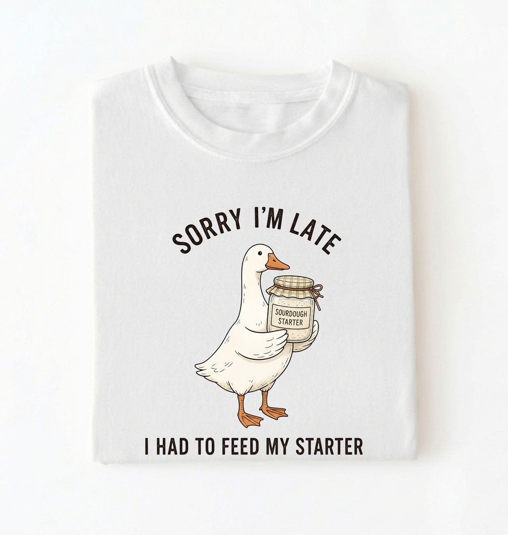 Flat lay of a white sorry im late i had to feed my starter shirt with goose and sourdough starter jar funny baker gift on a white background