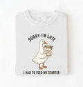 Flat lay of a white sorry im late i had to feed my starter shirt with goose and sourdough starter jar funny baker gift on a white background