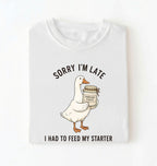 Flat lay of a white sorry im late i had to feed my starter shirt with goose and sourdough starter jar funny baker gift on a white background