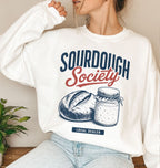 A woman wearing a white crewneck sweatshirt with a 'Sourdough Society' graphic print featuring navy blue and red lettering, an illustration of a sourdough loaf and a jar of flour, and a small 'Local Dealer' tag. She is wearing light-wash blue jeans in a cozy indoor setting with a small green plant on a wooden table to the side.