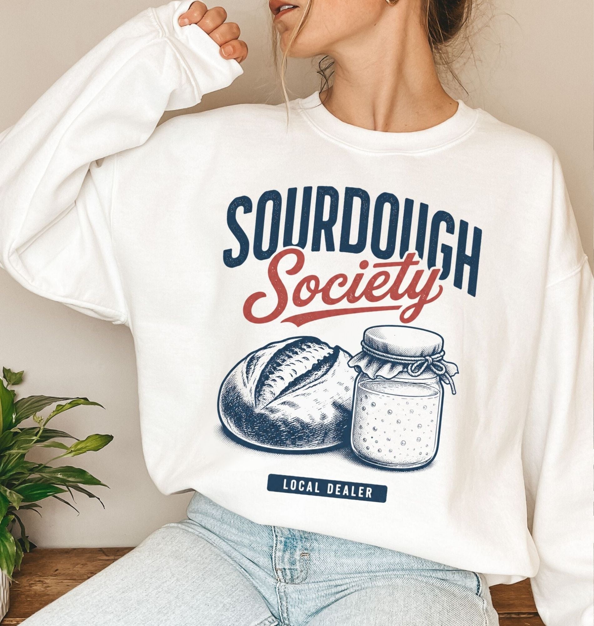 A woman wearing a white crewneck sweatshirt with a 'Sourdough Society' graphic print featuring navy blue and red lettering, an illustration of a sourdough loaf and a jar of flour, and a small 'Local Dealer' tag. She is wearing light-wash blue jeans in a cozy indoor setting with a small green plant on a wooden table to the side.
