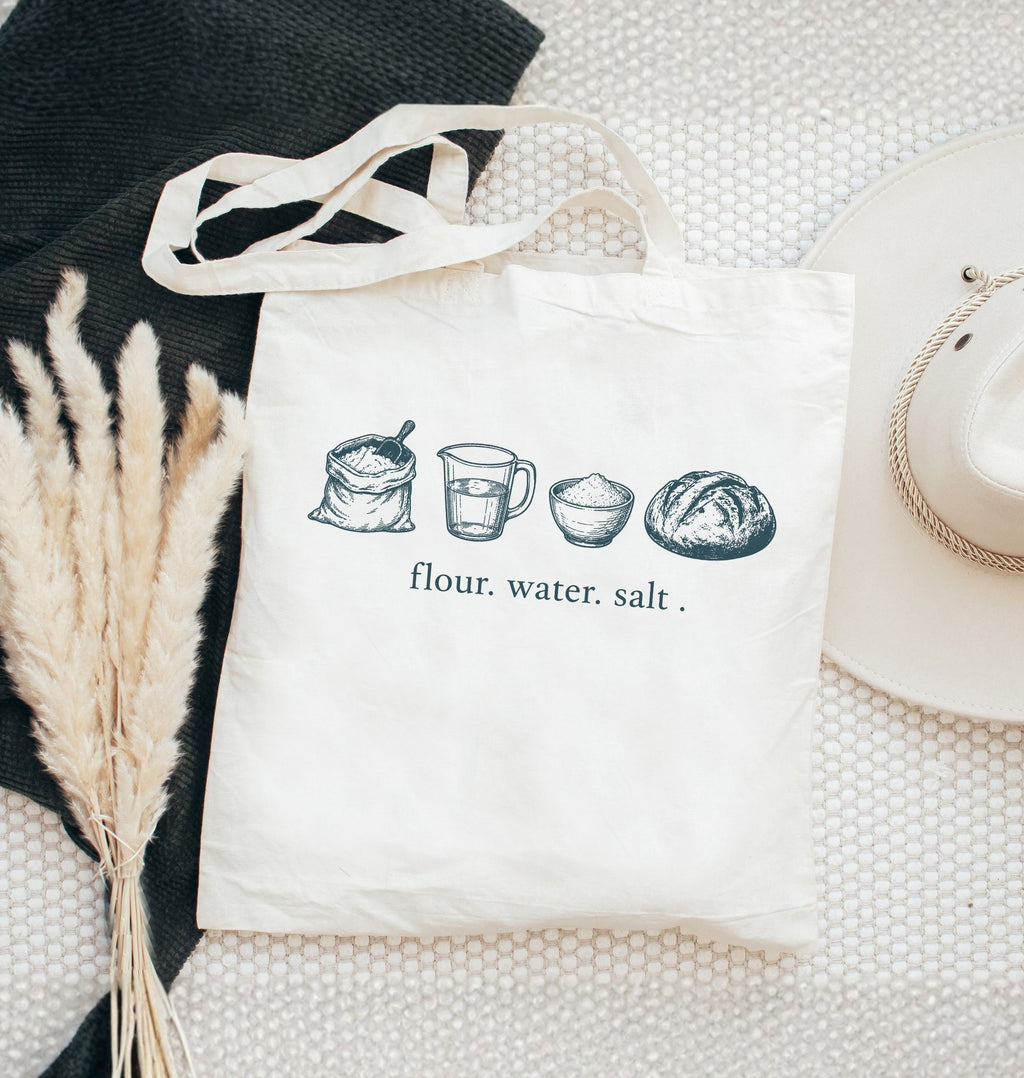 White tote bag with bread-making ingredients illustration and text on a textured surface.