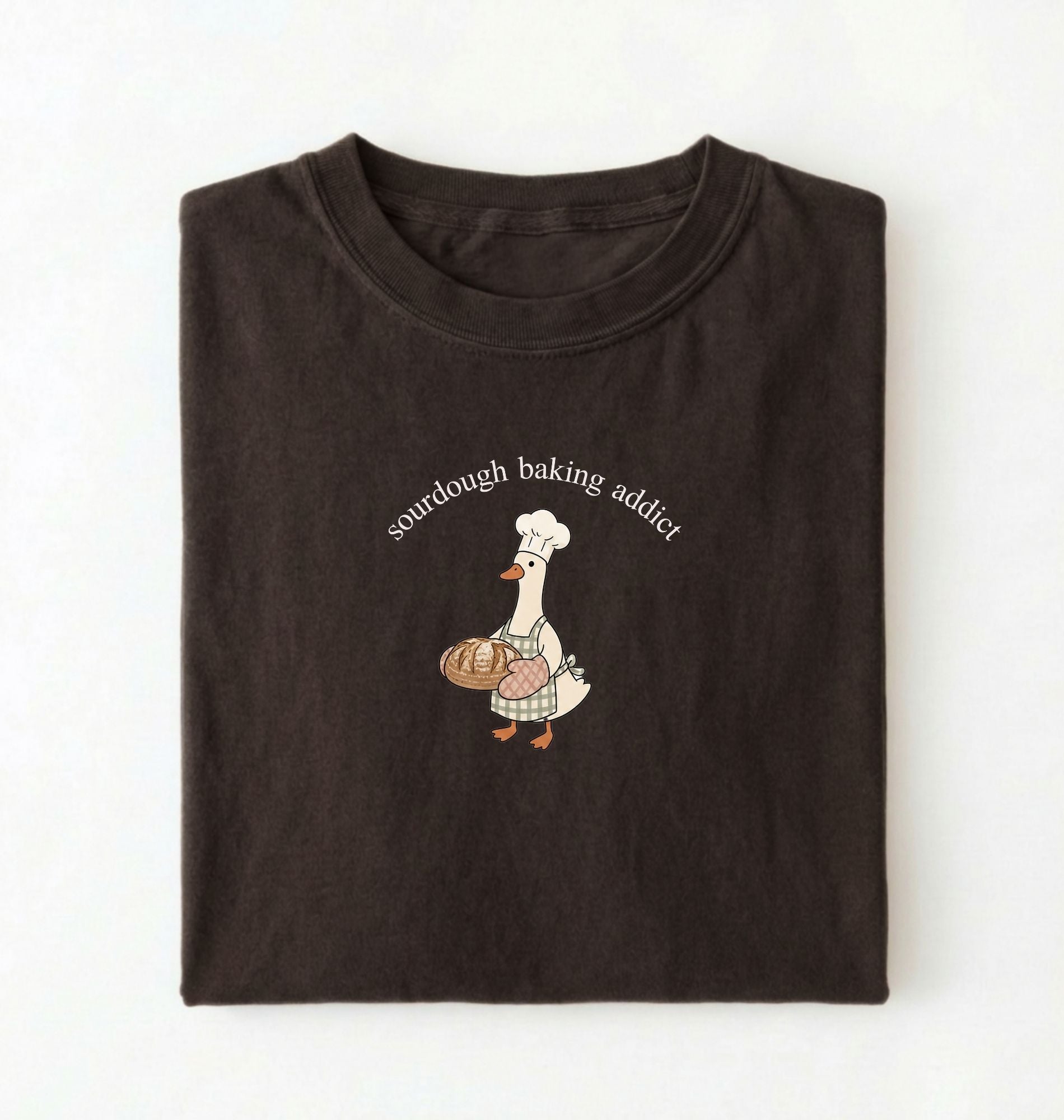 Flat lay of a dark chocolate Sourdough Baking Addict shirt with baker goose holding artisan bread for sourdough enthusiasts on a white background 