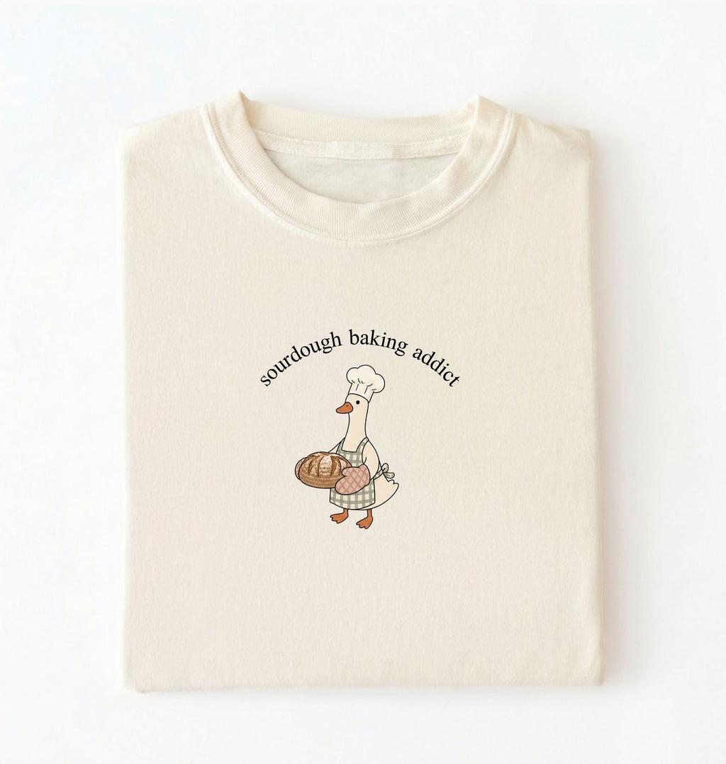 Flat lay of a ivory Sourdough Baking Addict shirt with baker goose holding artisan bread for sourdough enthusiasts on a white background 