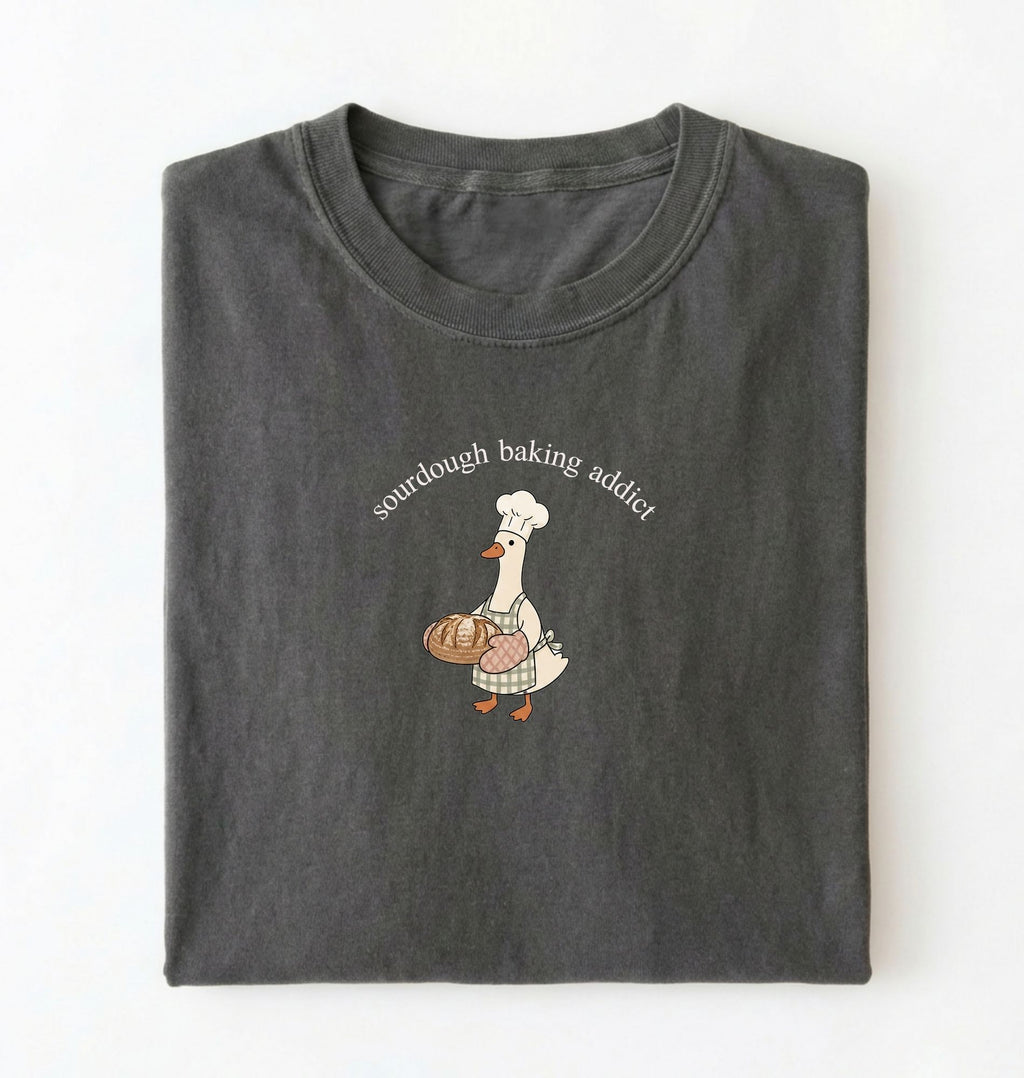 Flat lay of a dark grey Sourdough Baking Addict shirt with baker goose holding artisan bread for sourdough enthusiasts on a white background 