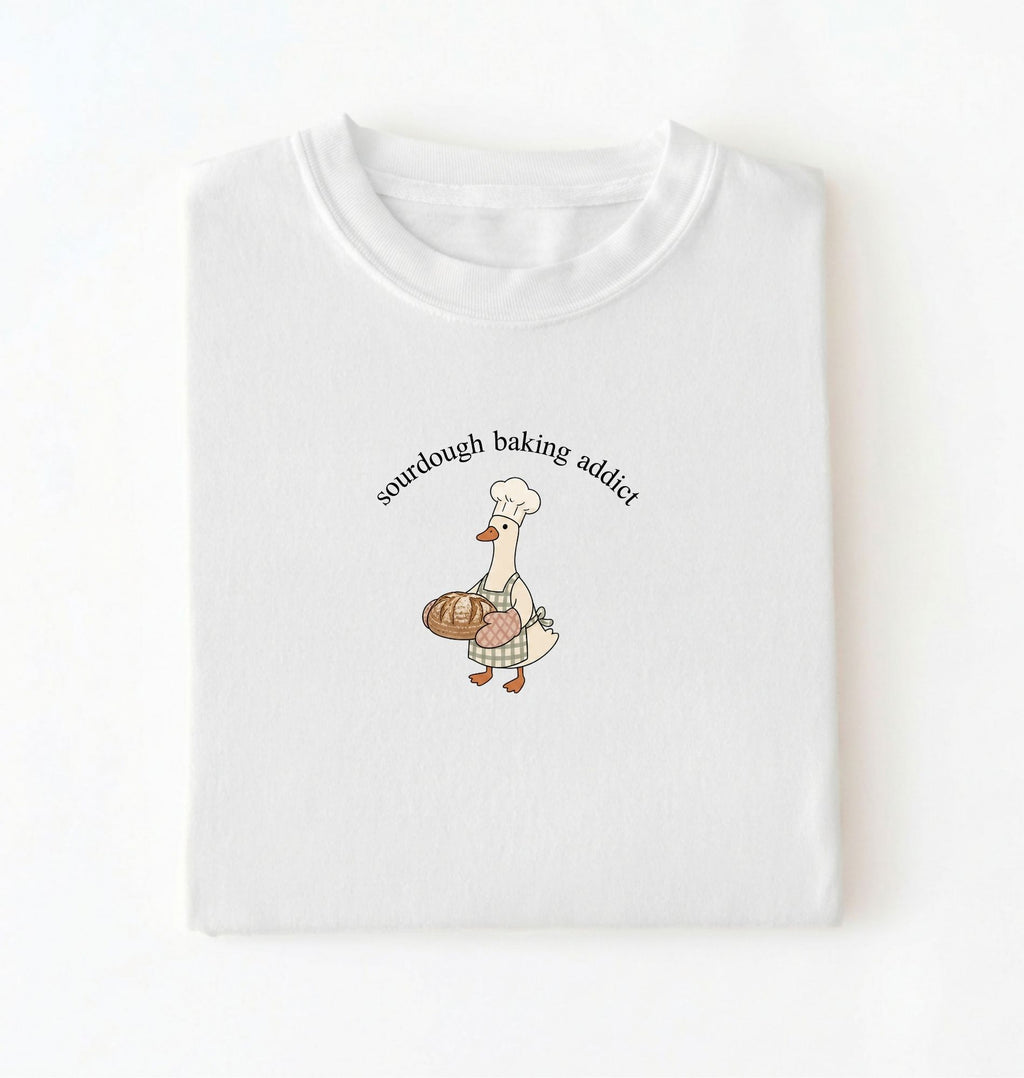 Flat lay of a white Sourdough Baking Addict shirt with baker goose holding artisan bread for sourdough enthusiasts on a white background 