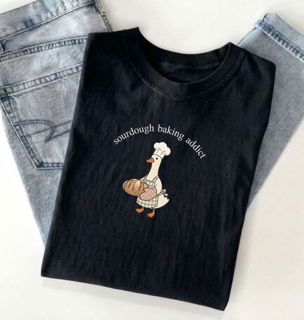 Black t-shirt with a graphic of a duck wearing a chef's hat and sourdough bread, text 'sourdough baking addict', on a light gray background.