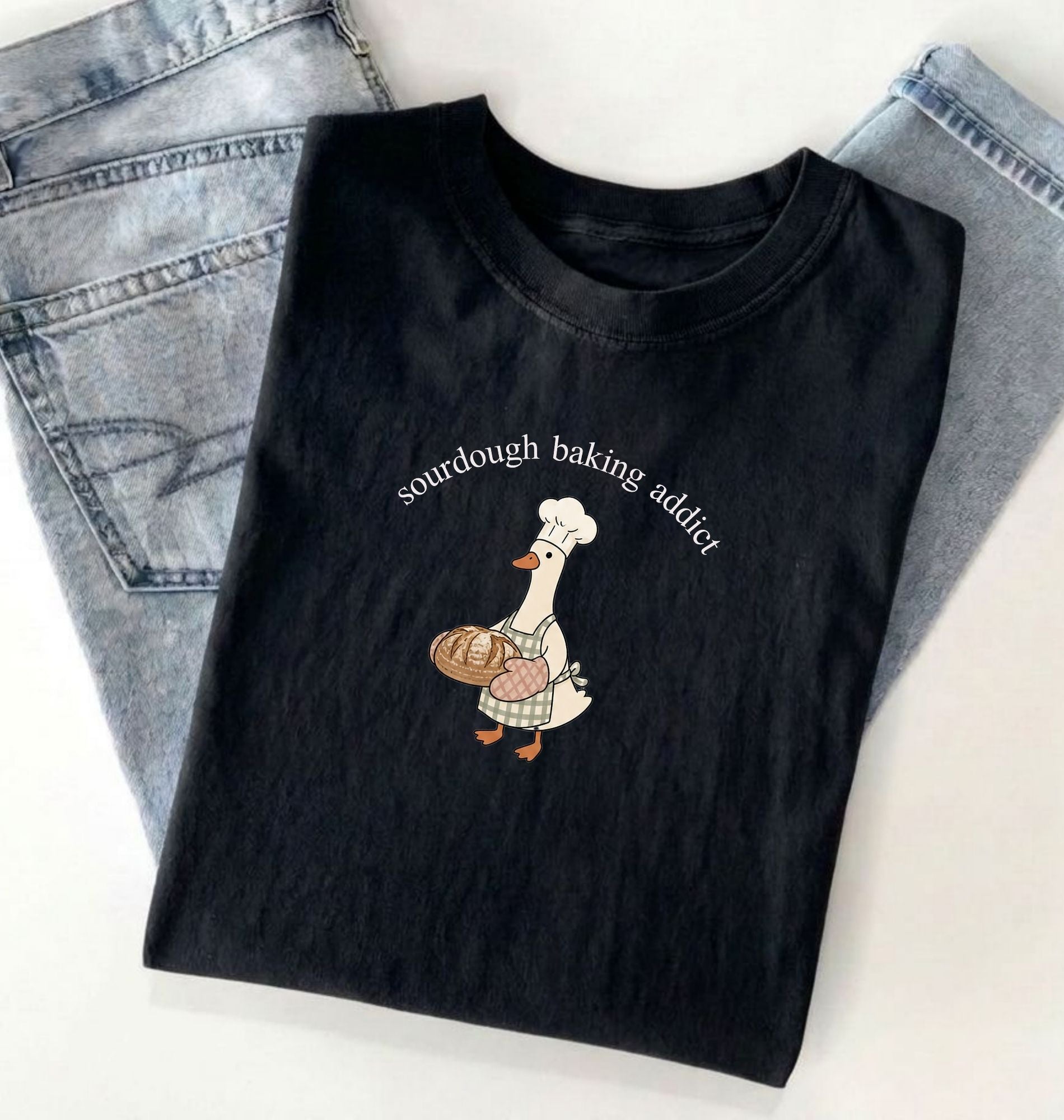 Black t-shirt with a graphic of a duck wearing a chef's hat and sourdough bread, text 'sourdough baking addict', on a light gray background.