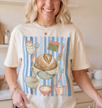 A woman wearing a cream-colored graphic t-shirt with a blue and brown illustration of baking ingredients and utensils, set against a warm kitchen background.