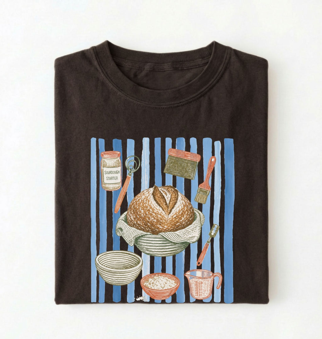 Flat lay of a brown Sourdough baking tools shirt featuring artisan bread, starter jar, banneton, and baking supplies on a white background 