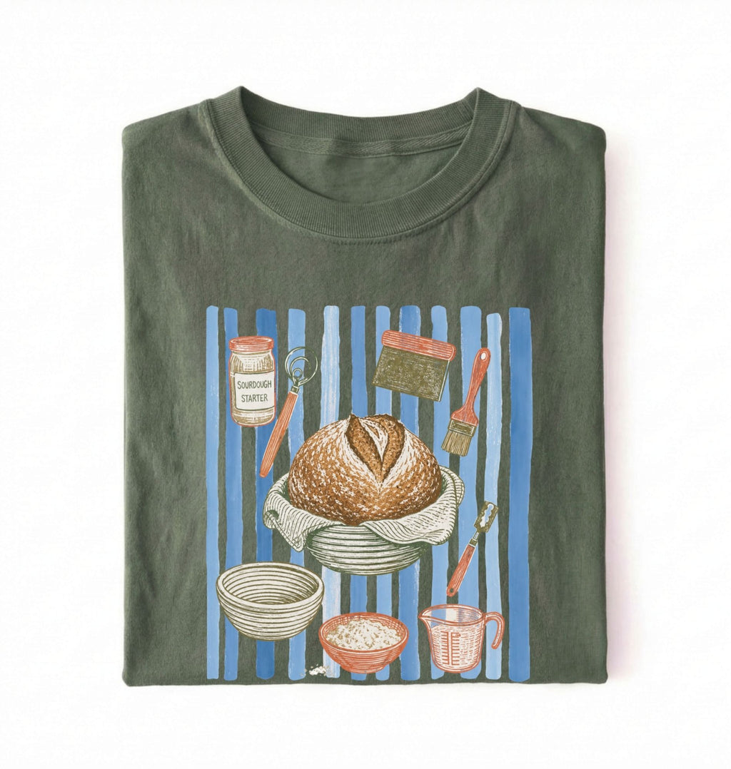 Flat lay of a green Sourdough baking tools shirt featuring artisan bread, starter jar, banneton, and baking supplies on a white background 