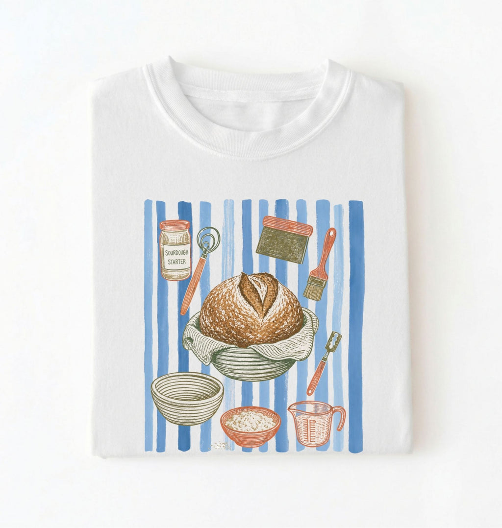 Flat lay of a brown Sourdough baking tools shirt featuring artisan bread, starter jar, banneton, and baking supplies on a white background 