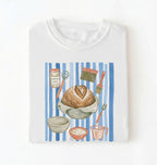 Flat lay of a brown Sourdough baking tools shirt featuring artisan bread, starter jar, banneton, and baking supplies on a white background 