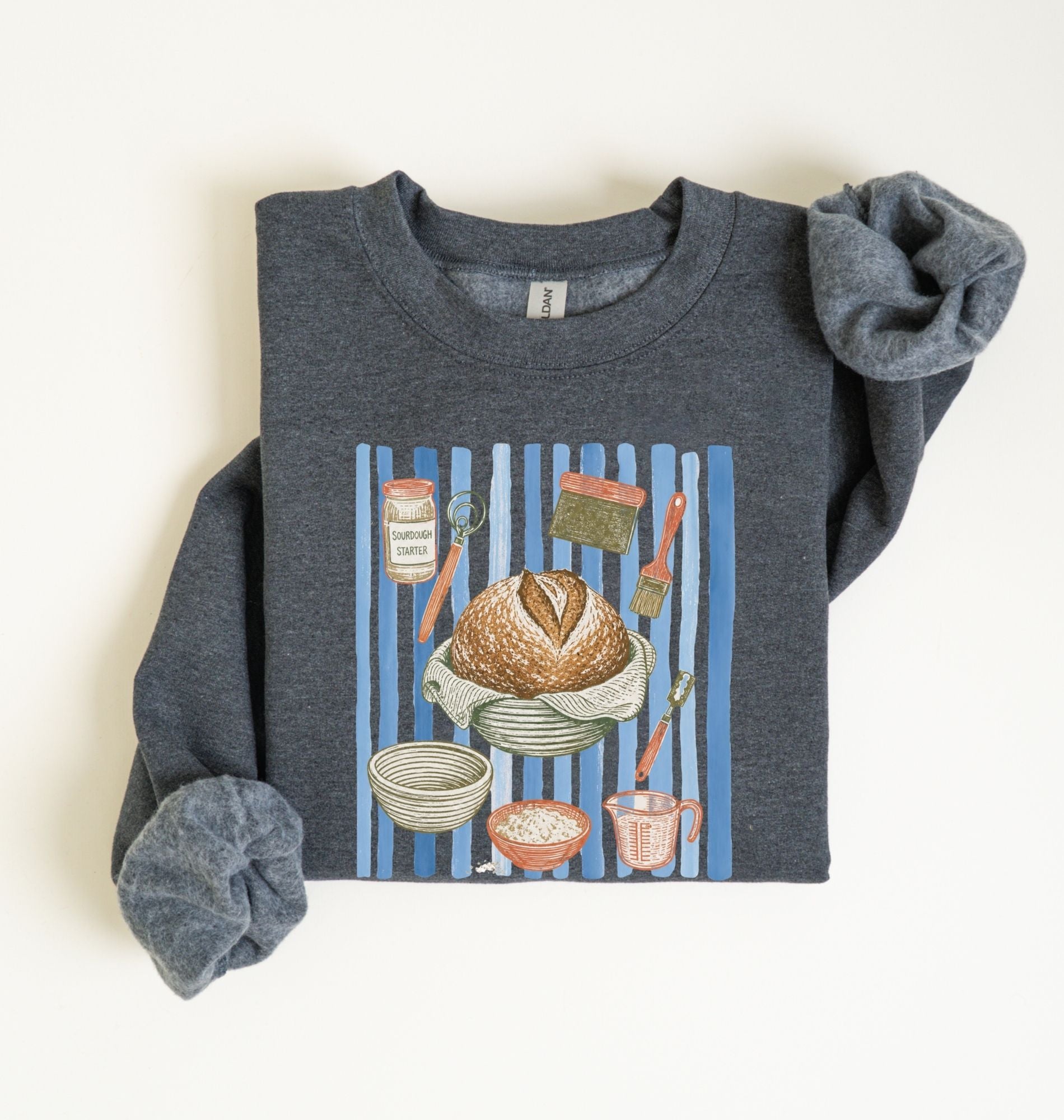 Flat lay of a dark heather Sourdough baking tools sweatshirt featuring artisan bread, starter jar, banneton, and baking supplies on a white background 