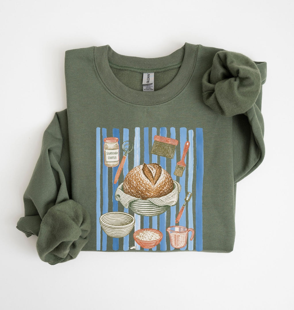 Flat lay of a green Sourdough baking tools sweatshirt featuring artisan bread, starter jar, banneton, and baking supplies on a white background 