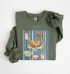 Flat lay of a green Sourdough baking tools sweatshirt featuring artisan bread, starter jar, banneton, and baking supplies on a white background 