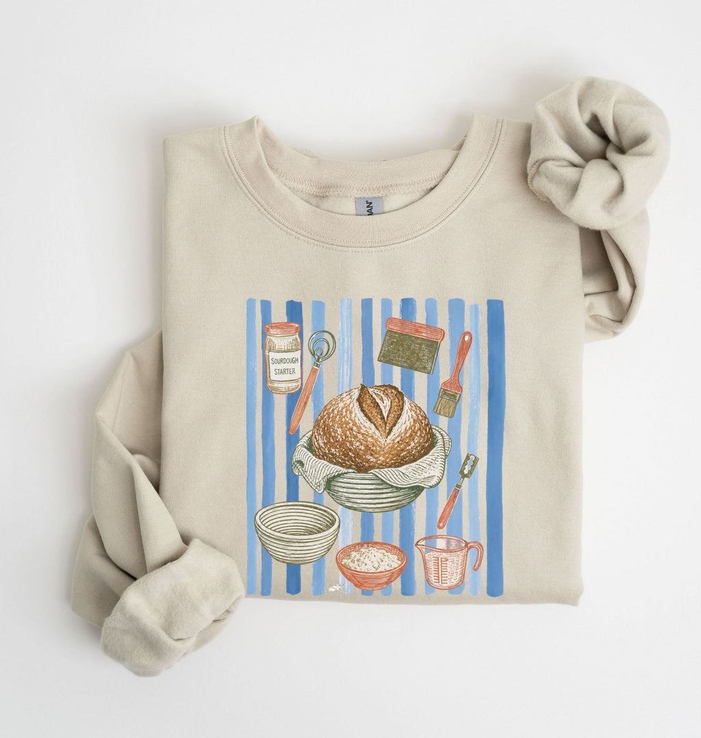 Flat lay of a sand Sourdough baking tools sweatshirt featuring artisan bread, starter jar, banneton, and baking supplies on a white background 