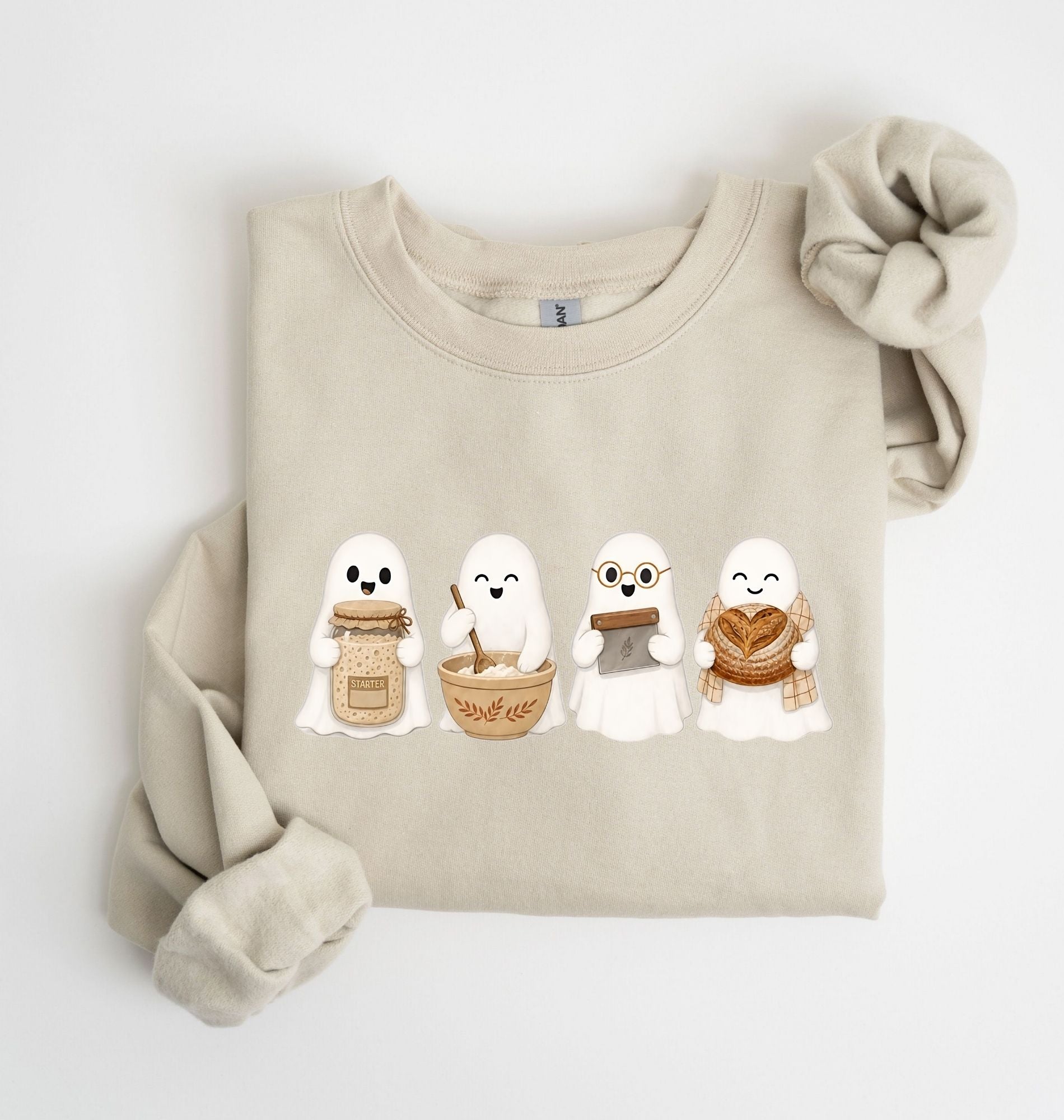 Flat lay of a sand sourdough sweatshirt featuring four cute baking ghosts with a starter jar, mixing bowl, bench scraper and bread loaf on a white background 