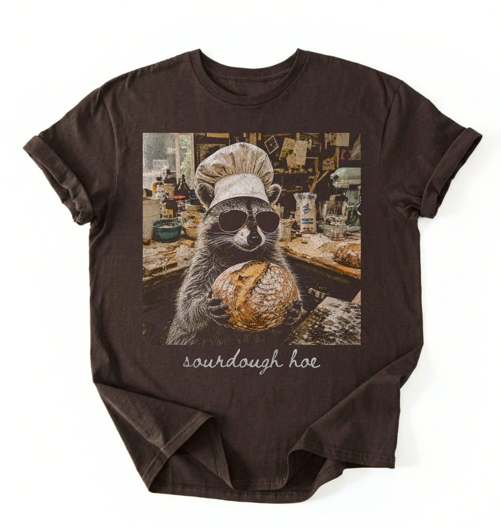 Flat lay of a sourdough hoe shirt brown funny raccoon baker t shirt sourdough bread lover gift on a white background 