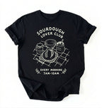 Flat lay of a black Sourdough Lover Club graphic shirt with artisan bread baking design for sourdough enthusiasts on a white background 