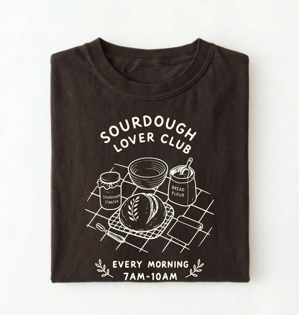 Flat lay of a brown  Sourdough Lover Club graphic shirt with artisan bread baking design for sourdough enthusiasts on a white background 
