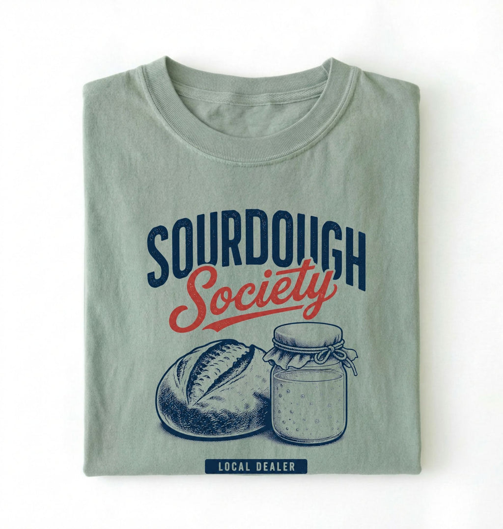 Flat lay of a bay sourdough society shirt with artisan bread and sourdough starter jar for bread bakers on a white background 