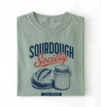 Flat lay of a bay sourdough society shirt with artisan bread and sourdough starter jar for bread bakers on a white background 