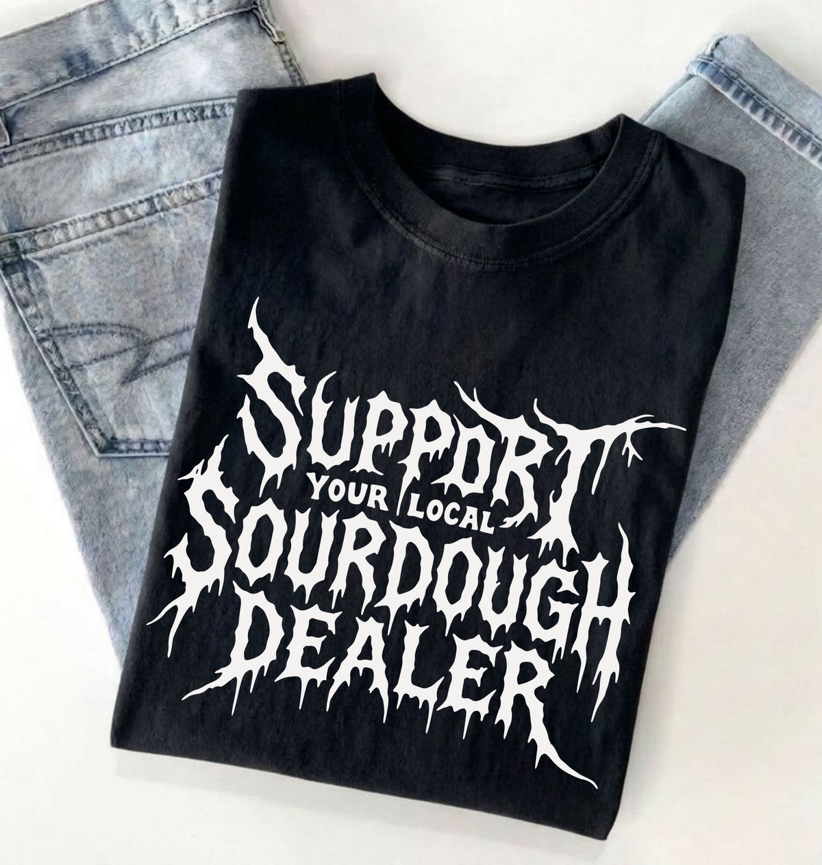 A black t-shirt with a white gothic-style graphic reading 'SUPPORT YOUR LOCAL SOURDOUGH DEALER' is laid flat on top of light-wash blue jeans against a white background.