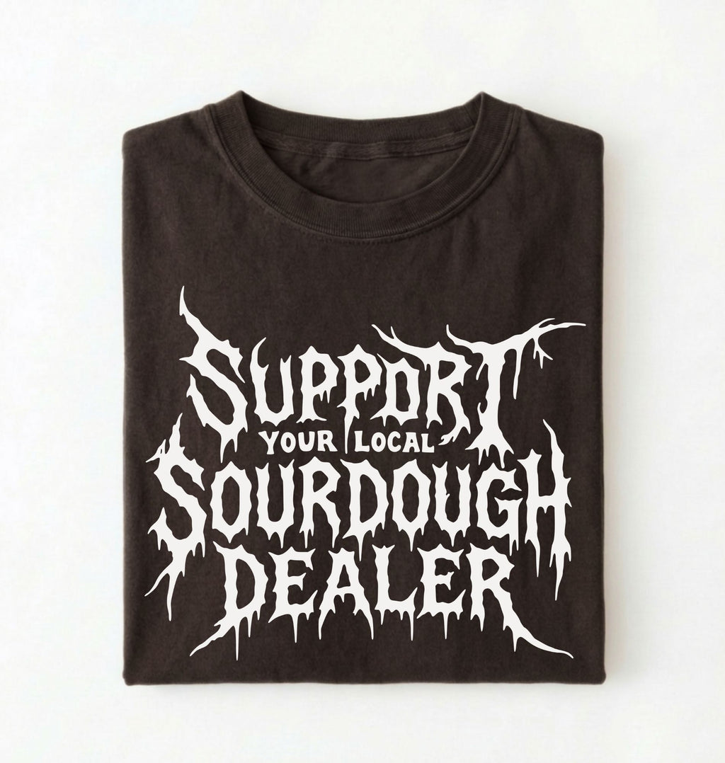 A folded black t-shirt with a white graphic print that reads 'SUPPORT YOUR LOCAL SOURDOUGH DEALER' in a stylized, dripping font, displayed against a plain white background.
