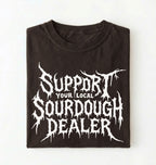 A folded black t-shirt with a white graphic print that reads 'SUPPORT YOUR LOCAL SOURDOUGH DEALER' in a stylized, dripping font, displayed against a plain white background.