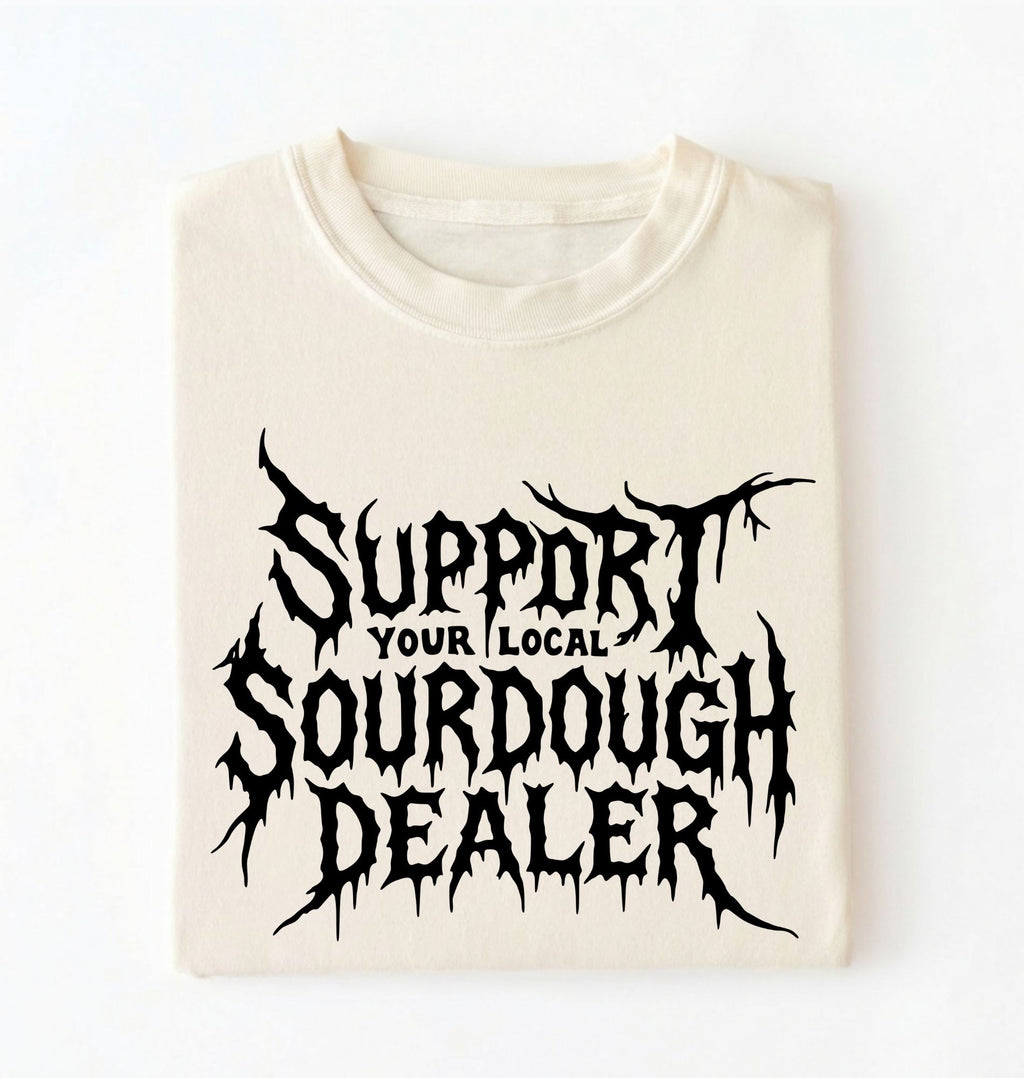 A folded cream-colored t-shirt with a black gothic-style graphic that reads 'SUPPORT YOUR LOCAL SOURDOUGH DEALER' on a plain white background.