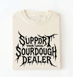 A folded cream-colored t-shirt with a black gothic-style graphic that reads 'SUPPORT YOUR LOCAL SOURDOUGH DEALER' on a plain white background.