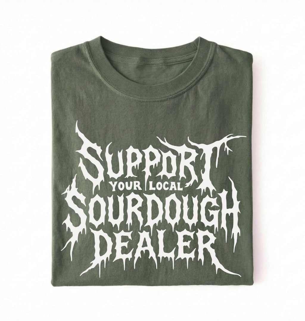 A folded dark olive green t-shirt with a white graphic print that reads 'SUPPORT YOUR LOCAL SOURDOUGH DEALER' in a stylized, dripping font, displayed against a plain white background.