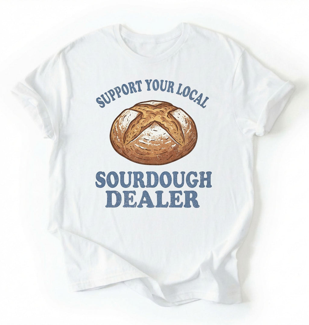 Flat lay of a Support Your Local Sourdough Dealer shirt with artisan sourdough bread graphic on white tee on a white background 