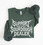A dark green crewneck sweatshirt with 'Support Your Local Sourdough Dealer' printed in a white, stylized horror font on the front, laid flat on a white background with the sleeves folded.