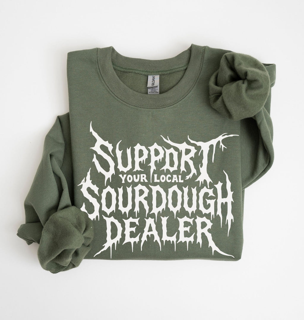An olive green crewneck sweatshirt with rolled-up sleeves, featuring a white graphic print that reads 'SUPPORT YOUR LOCAL SOURDOUGH DEALER' in a stylized font, laid flat on a white background.