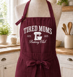 A burgundy kitchen apron with a white skeleton graphic and the text 'HAVING A MENTAL BAKE DOWN' displayed on a mannequin torso in a bright kitchen setting.