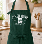A dark green kitchen apron with white text reading 'TIRED MOMS Baking Club' and a mixer graphic, displayed on a mannequin in a bright kitchen setting.