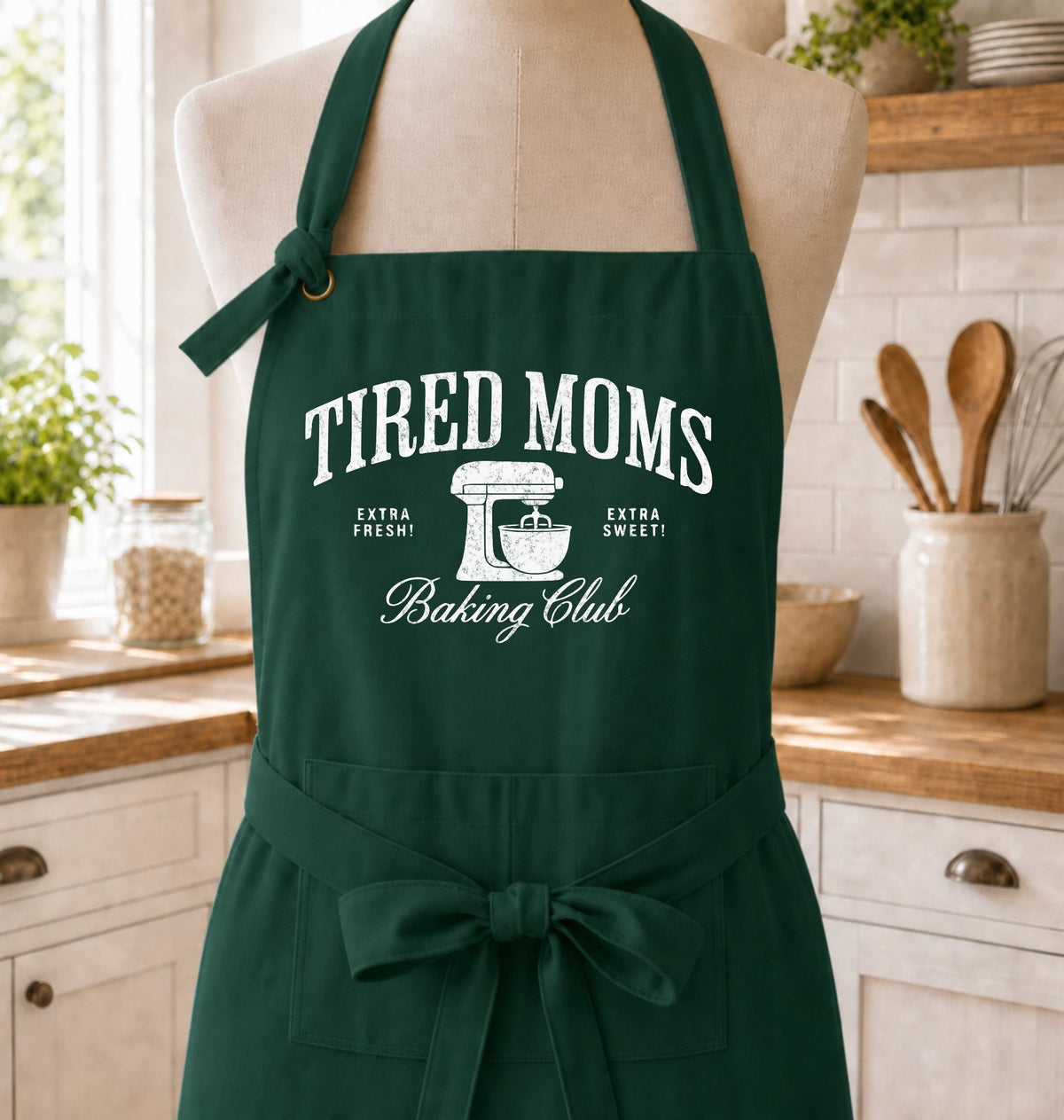 A dark green kitchen apron with white text reading 'TIRED MOMS Baking Club' and a mixer graphic, displayed on a mannequin in a bright kitchen setting.