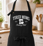 A black apron displayed on a beige mannequin torso in a bright kitchen setting. The apron features white printed text reading 'TIRED MOMS Baking Club' with a stand mixer graphic and smaller phrases 'EXTRA FRESH!' and 'EXTRA SWEET!'.