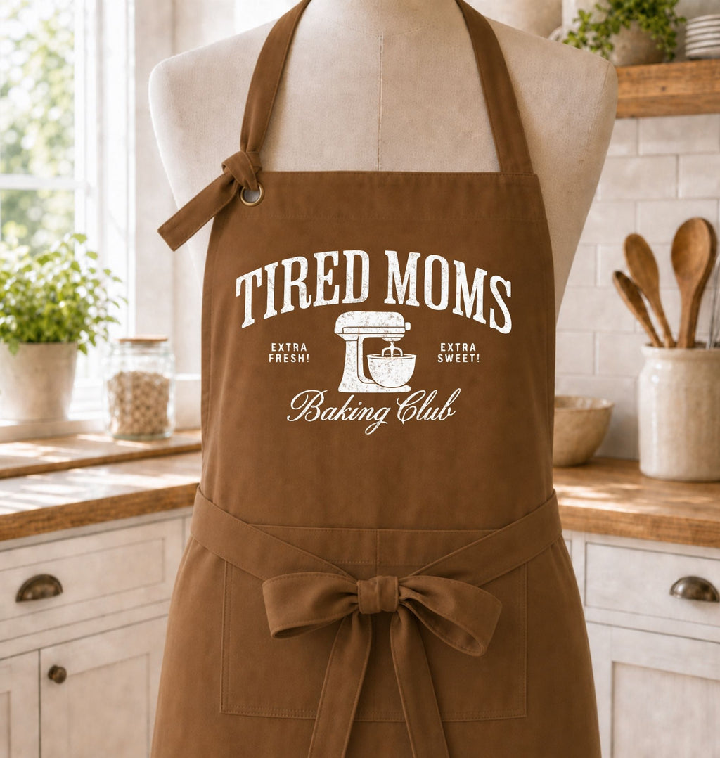 A brown apron with white printed text reading 'TIRED MOMS Baking Club' and a mixer graphic, displayed on a mannequin in a bright kitchen setting.