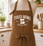 A brown apron with white printed text reading 'TIRED MOMS Baking Club' and a mixer graphic, displayed on a mannequin in a bright kitchen setting.