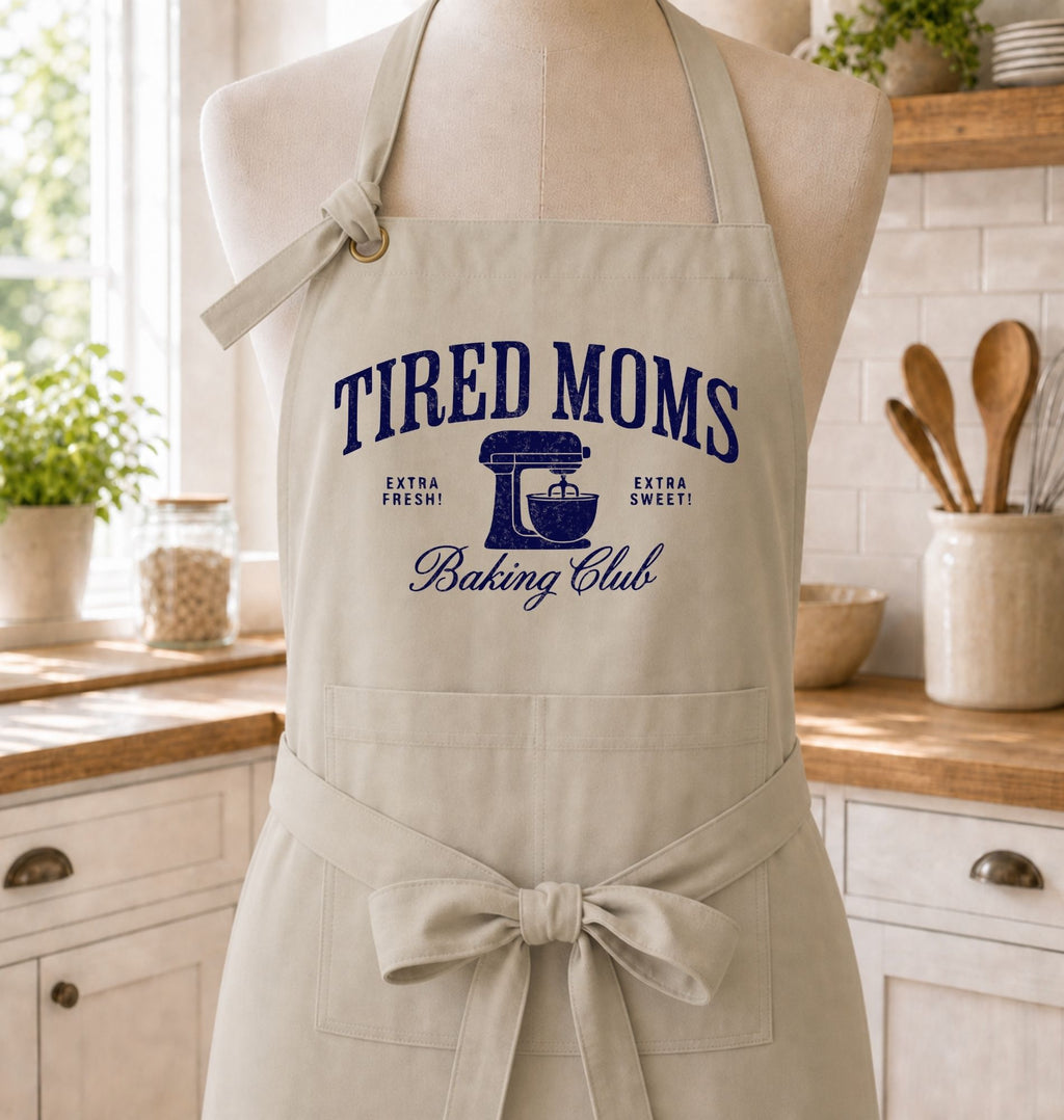 A beige apron with a navy blue graphic reading 'TIRED MOMS Baking Club' is displayed on a mannequin in a bright kitchen setting. The apron features a neck strap, waist tie, and front pockets.