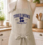 A beige apron with a navy blue graphic reading 'TIRED MOMS Baking Club' is displayed on a mannequin in a bright kitchen setting. The apron features a neck strap, waist tie, and front pockets.