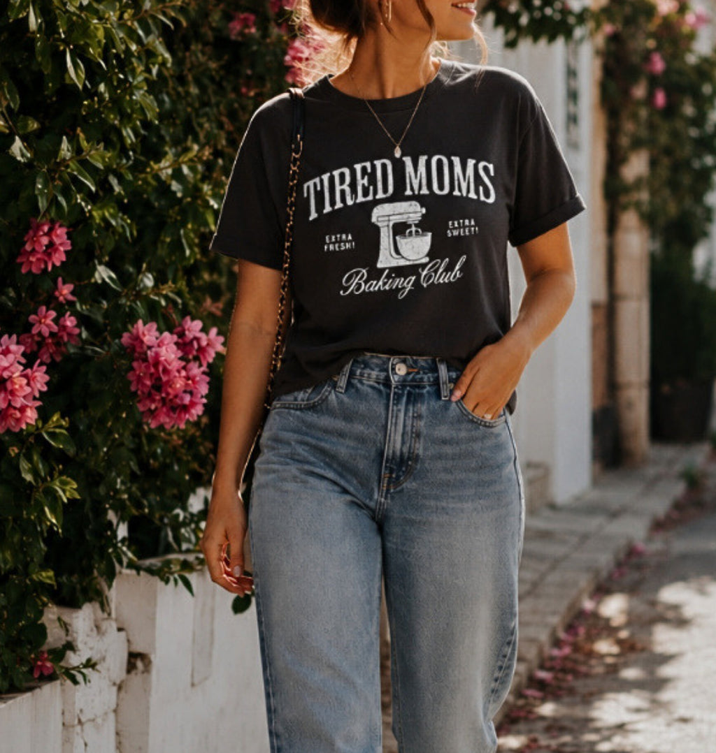 A woman wearing a black graphic t-shirt that reads 'TIRED MOMS Baking Club' in white lettering, paired with light-wash blue jeans, standing outdoors on a sunlit sidewalk next to a white wall and green foliage with pink flowers.