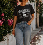 A woman wearing a black graphic t-shirt that reads 'TIRED MOMS Baking Club' in white lettering, paired with light-wash blue jeans, standing outdoors on a sunlit sidewalk next to a white wall and green foliage with pink flowers.